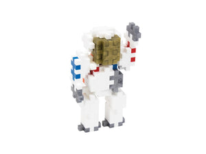 A small astronaut toy figure built from interlocking blocks, standing on a plain white background. The figure is mostly white with gray limbs and feet, a tan helmet visor, blue accents on one shoulder, and red accents on the chest.
